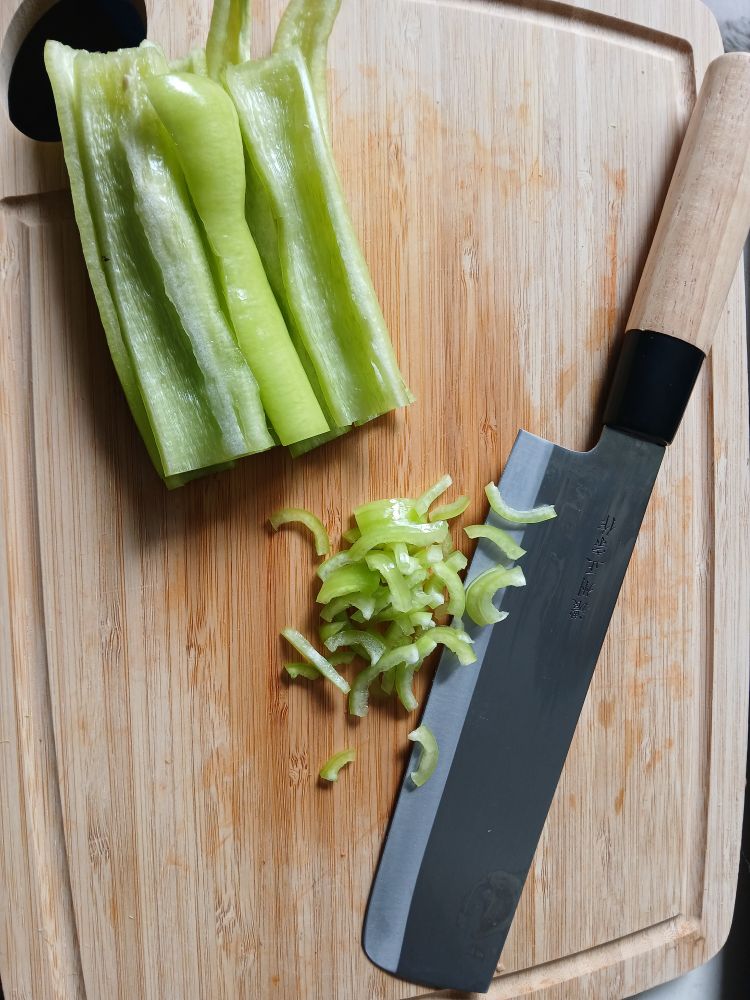 Ein Schneidbrett aus Bambus mit einem Nakiri-Messer, breiten Längsstreifen von grünen Paprika und von diesen geschnittene hauchfeine Querstreifen.