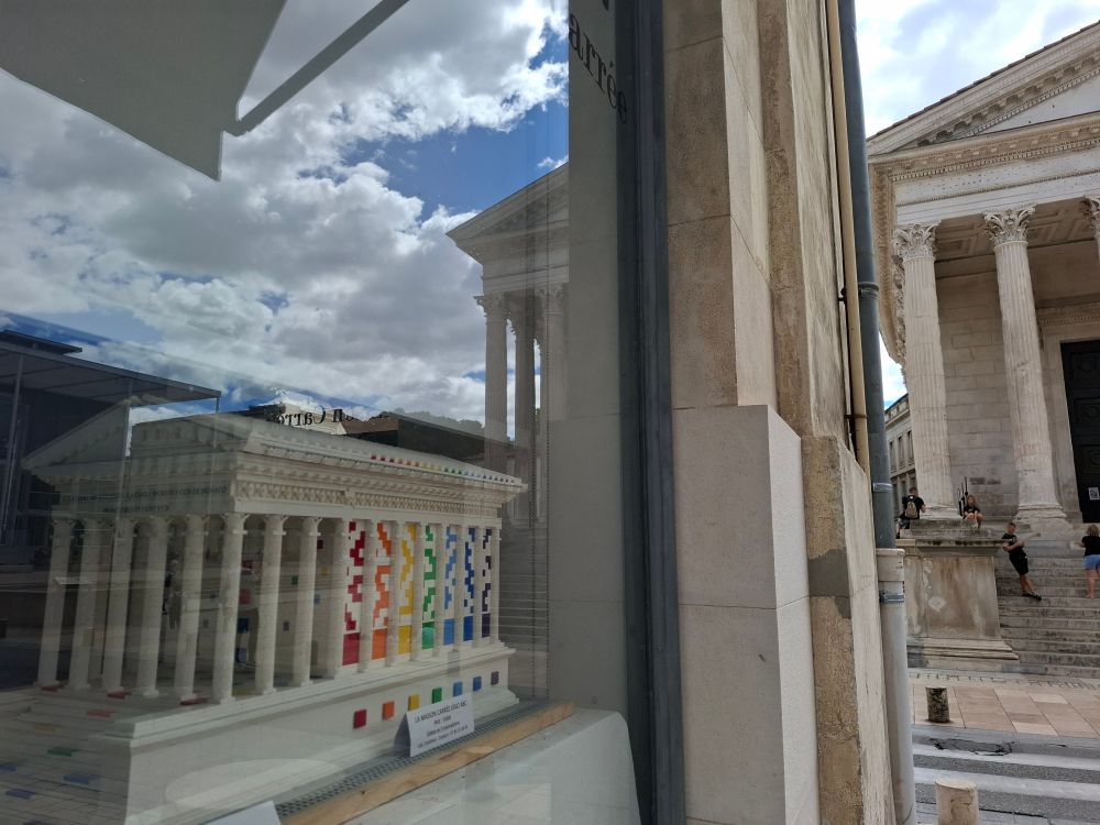 Maison carrée à Nîmes se reflétant dans une vitrine derrière laquelle on voit une maquette LEGO multicolore de la Maison carrée.