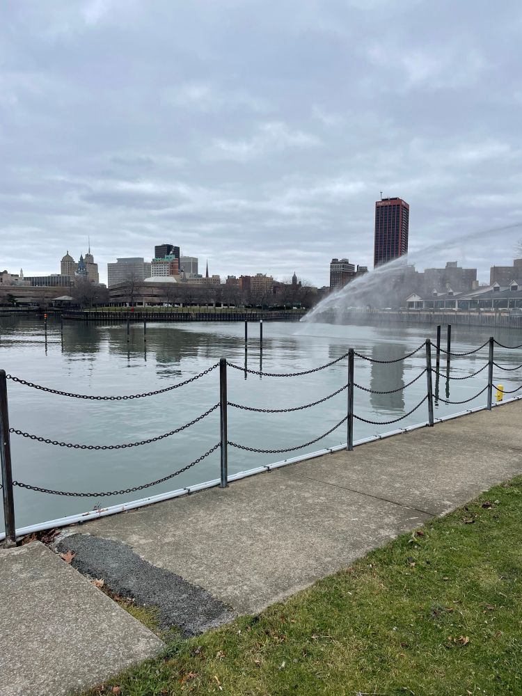 A sidewalk with a chain fence in front of water; beyond is the city of Buffalo skyline. A dramatic arc of water shoots in from the right side into the harbor.