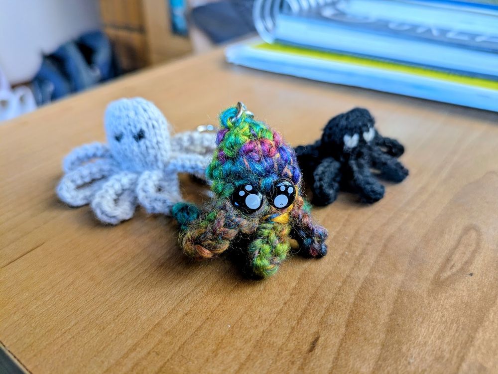 Photo of three small crocheted octopuses on a desk. The one in the center is beautifull handmade with rainbow yarn and two adorable button eyes, and a little metal link on top connecting it to a hidden keychain. It is flanked by simpler machine-made octopus finger puppets in gray and black.