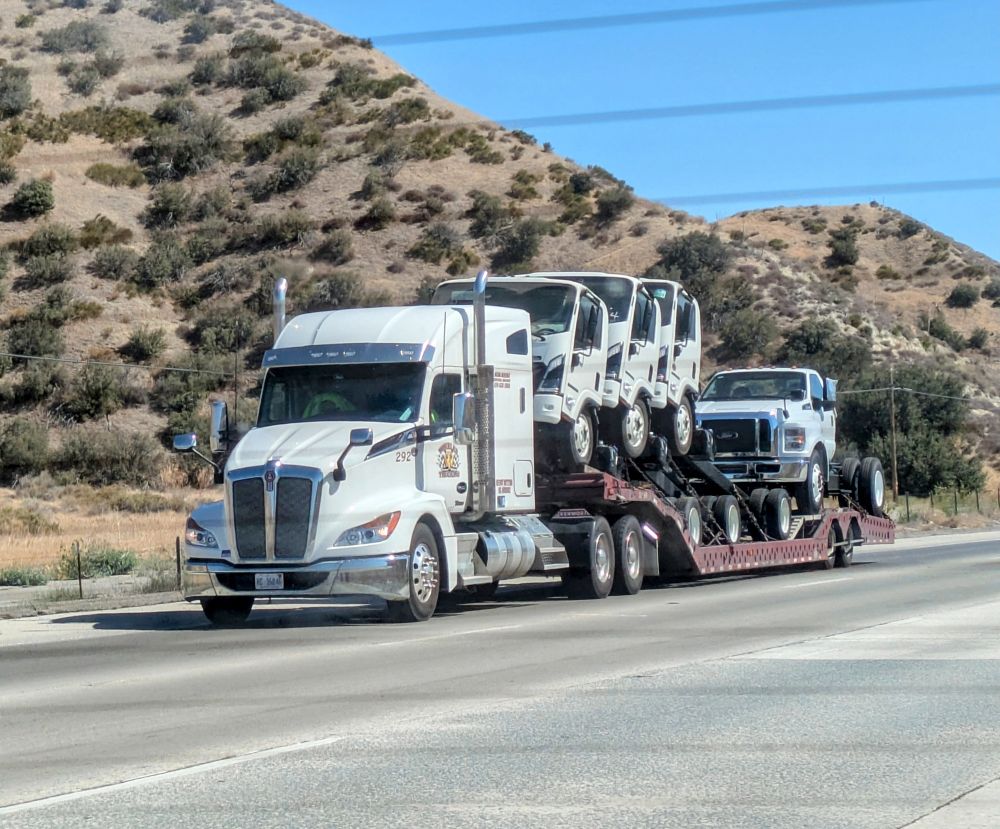 Photo of a white truck (possibly a Snowy Peterbilt?) in motion, carrying four small trucks on its bed (dorsal side).