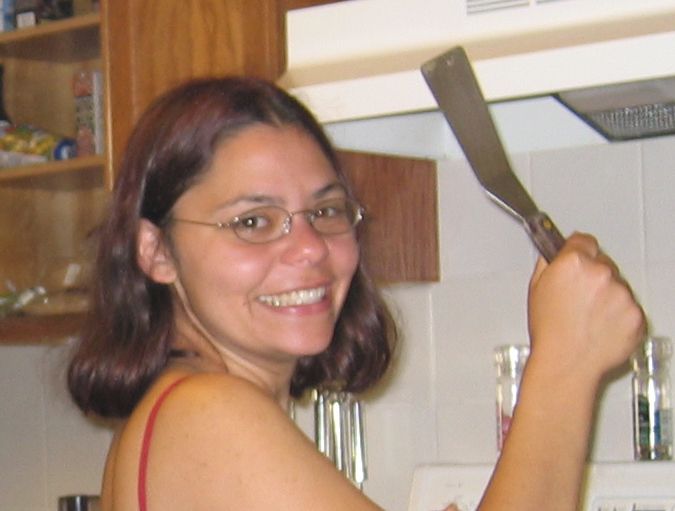 A blurry photo crop of me (Danna Staaf) grinning at the camera and brandishing a spatula. I think I was making pancakes with leftover vegetables? My hair is dark brown and shoulder length (with faint red highlights left over, like the vegetables, from something experimental--grad school was Like That) and I'm wearing glasses with oval lenses.