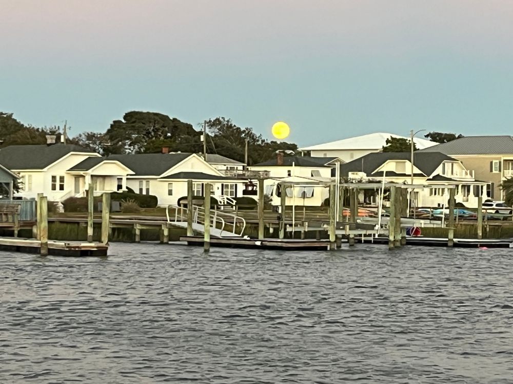 View across the creek to a shore lined with empty docks, houses behind, trees behind the houses, and a big yellow moon just over the trees