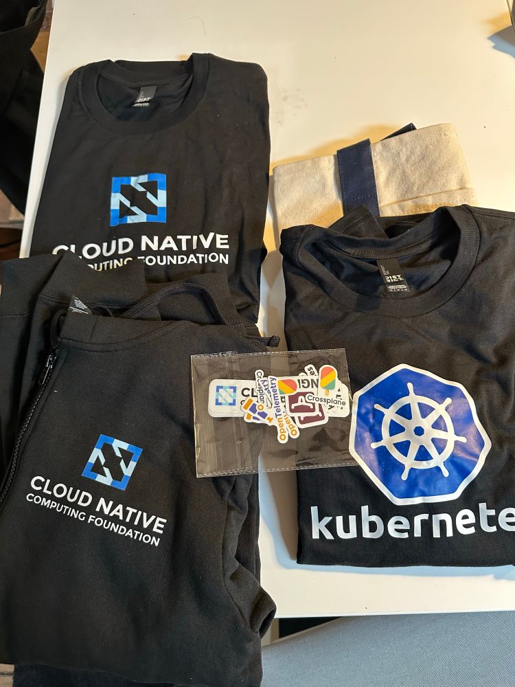A picture of CNCF and Kubernetes merch (two graphic tees, a sweat jacket and some project stickers)