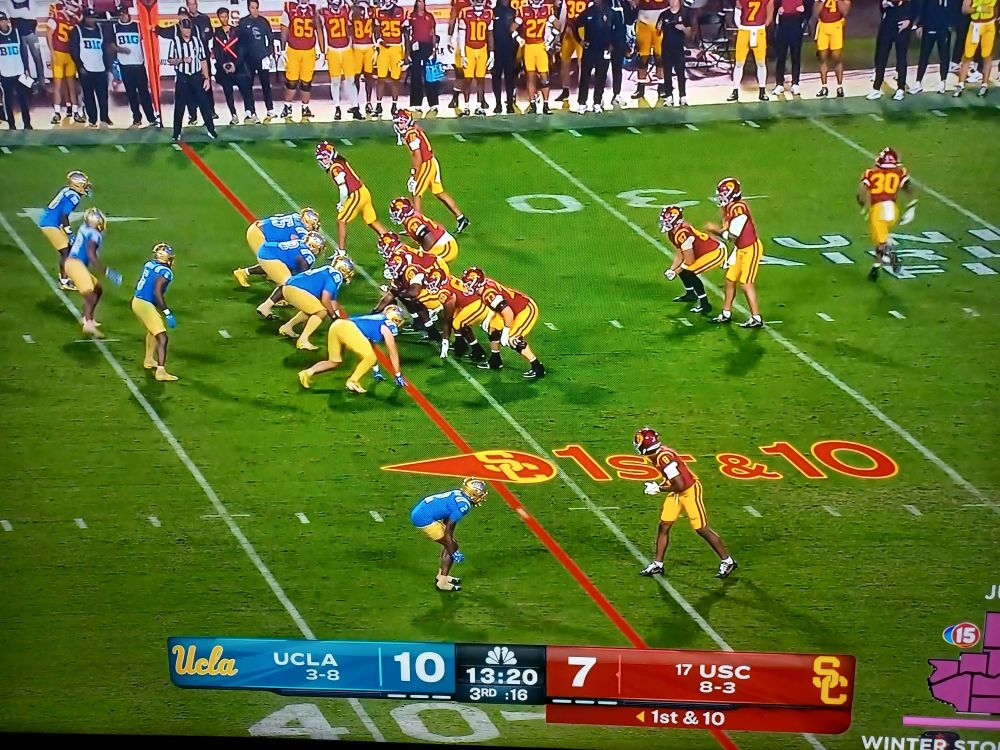 Image of a television screen showing two football teams lined up on a green football field just prior to a play from scrimmage. The team on offense, USC, wears vivid dark red jerseys and helmets and gold/yellow pants, while the team on defense, UCLA, wears bright sky-blue jerseys and a slightly duller gold on their pants and shiny gold helmets. 