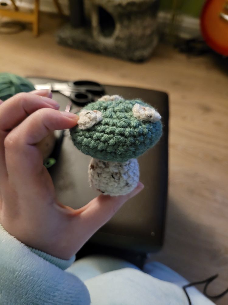 A small green crochet mushroom with an off-white stalk and three dots of the same color on the cap - being gently squeezed 