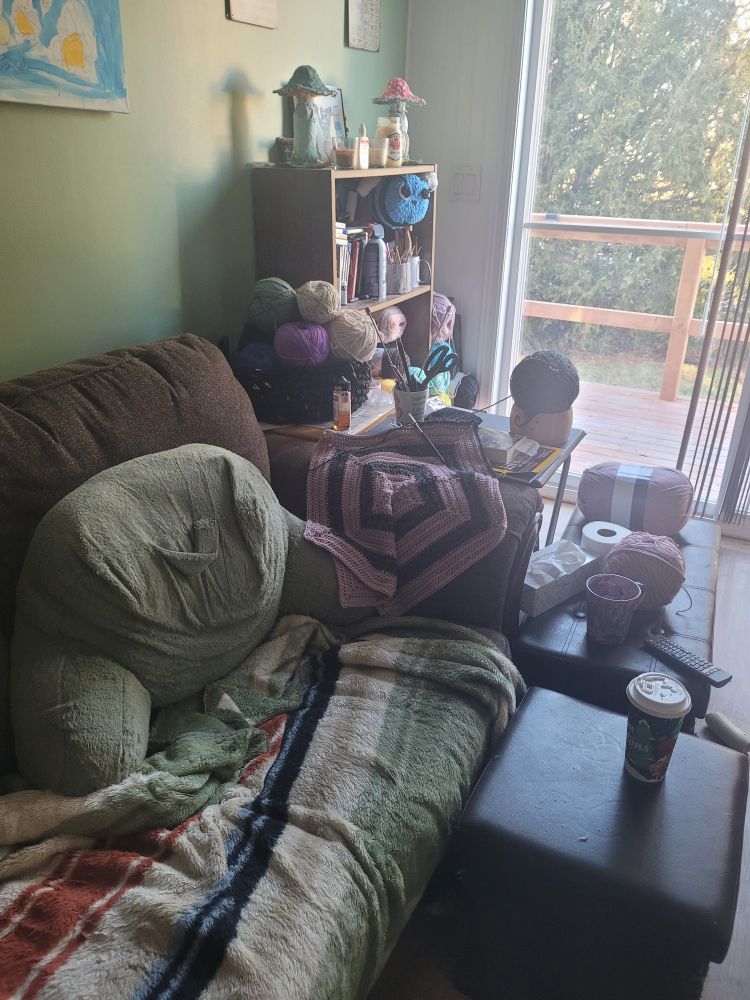 A cozy corner of the couch next to a glass patio door. A crochet work in progress is on the arm of the couch, and there is yarn surrounding the side of the couch. There is a back rest pillow and a fleece blanket where I sit, a coffee in front of me.