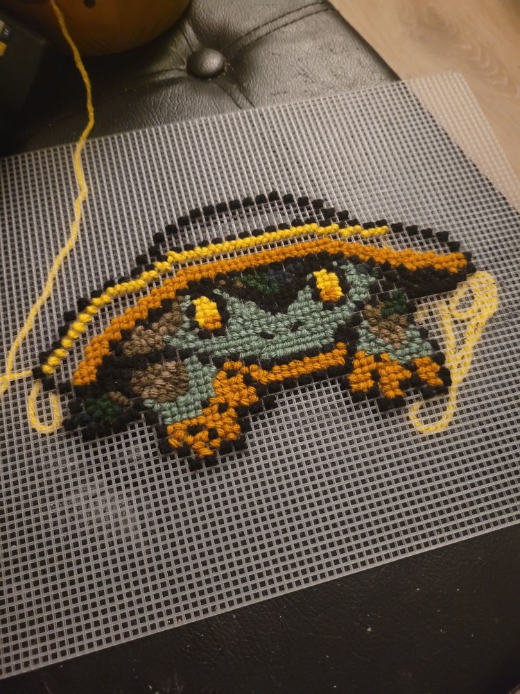 A cross stitch of a green and brown frog with orange accents wearing a hat. This is done with plastic mesh rather than cloth and stitched with a yarn needle and medium weight yarn. 