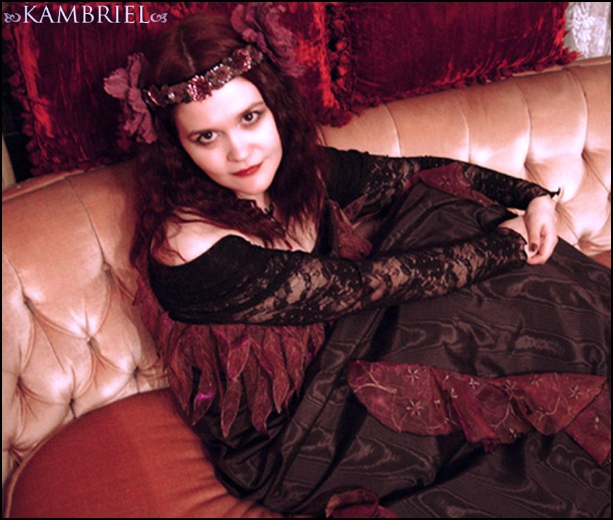 Portrait of Kambriel looking upwards, wearing an Art Nouveau floral headdress & black dress with luminous embroidered accents of her own making ~ sitting on an antique apricot velvet Victorian sofa that's backed with a pair of ruby crushed velvet pillows. (photo: Curse)