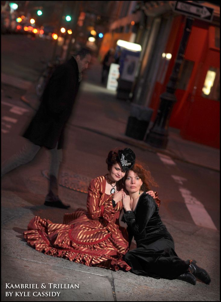 Evening portrait of Kambriel (in top hat, and formal red/gold striped bustle gown) & Trillian Stars (in black velvet) sitting on NYC asphalt as a man casually strolls by. Photo: Kyle Cassidy