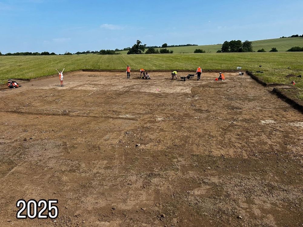 A small group of students and staff stand in a huge open trench in the middle of a field. The sky is blue!