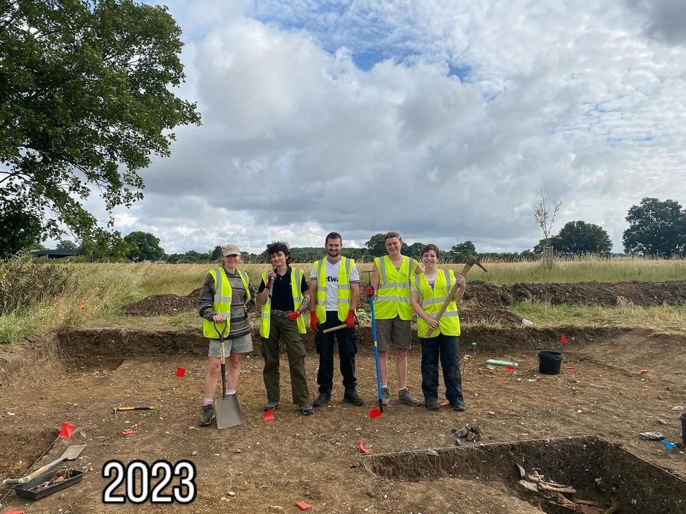 A team of students in yellow hi vis vests stand in an open trench holding tools. The sky is cloudy and they are surrounded by fields.