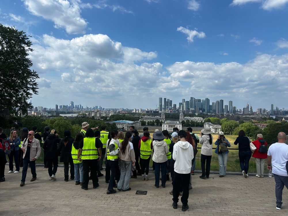 A group of students and members of the public stand at the view point in Greenwich Royal Park overlooking the Naval College and the Thames and City of London in the background. 