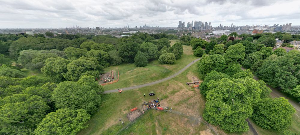 The Greenwich Archaeology Project from above looking North over London.  The green park and trees are in the foreground, with the trenches in the centre of the image, with the high rise buildings of the city of London in the background. You can just make out the River Thames as well. Photo by Pete Barry.