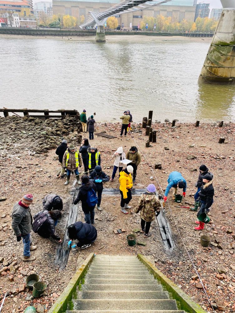 Students and staff from the UCL Institute of Archaeology on the foreshore or beach of the Thames working at the bottom of a staircase. They are cleaning a former 17th century staircase.  Behind them is the Thames and beyond that is the Tate Modern. 