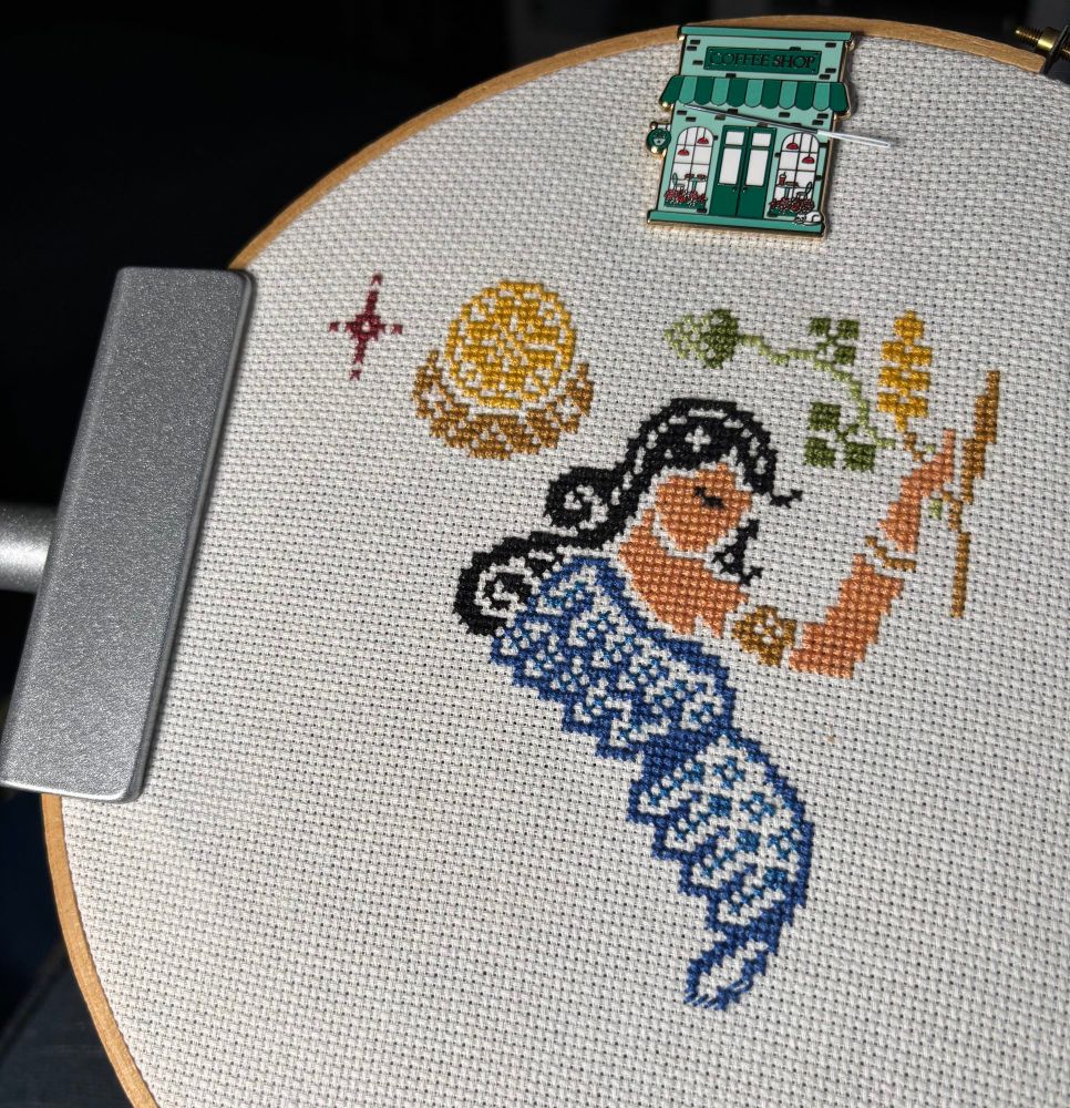 Cross stitch of Sumerian goddess of beer, Ninkasi, with long black hair, a blue dress, holding hops in her hand. 