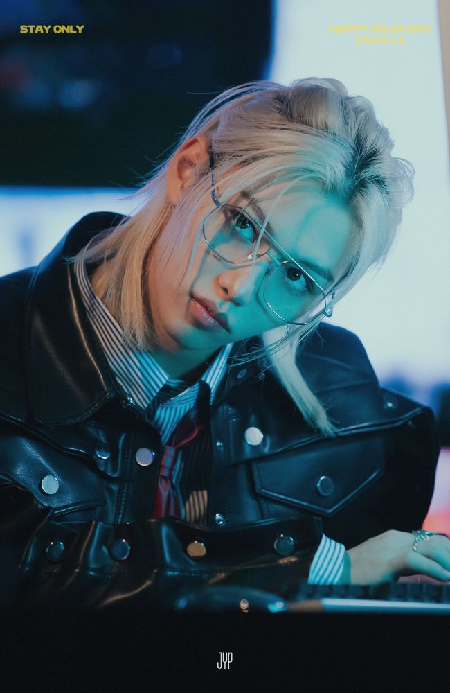He is wearing old school silver rimmed glasses, his hair half up, a black biker leather jacket, a striped Button down shirt and a red tie. His head is in an angle.