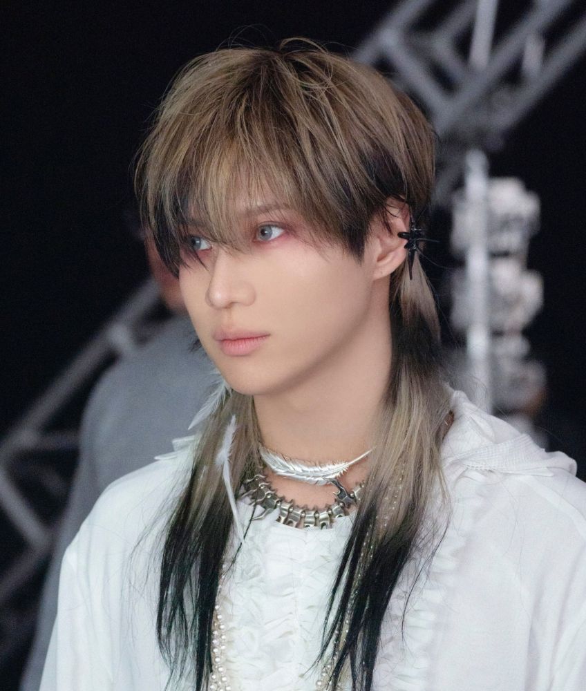 his hair is dark blonde with black stripes, he is wearing some extensions, blue contact lenses and a pretty pinkish eye make up. he is looking intensely at something in front of him. he has a feather chocker and is wearing a white frilly long sleeved blouse.