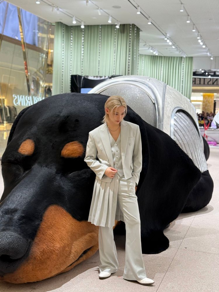 He is standing in front of the enormous tamburins dog. He is wearing a light blue suit with vest and a half skirt. He is looking down.