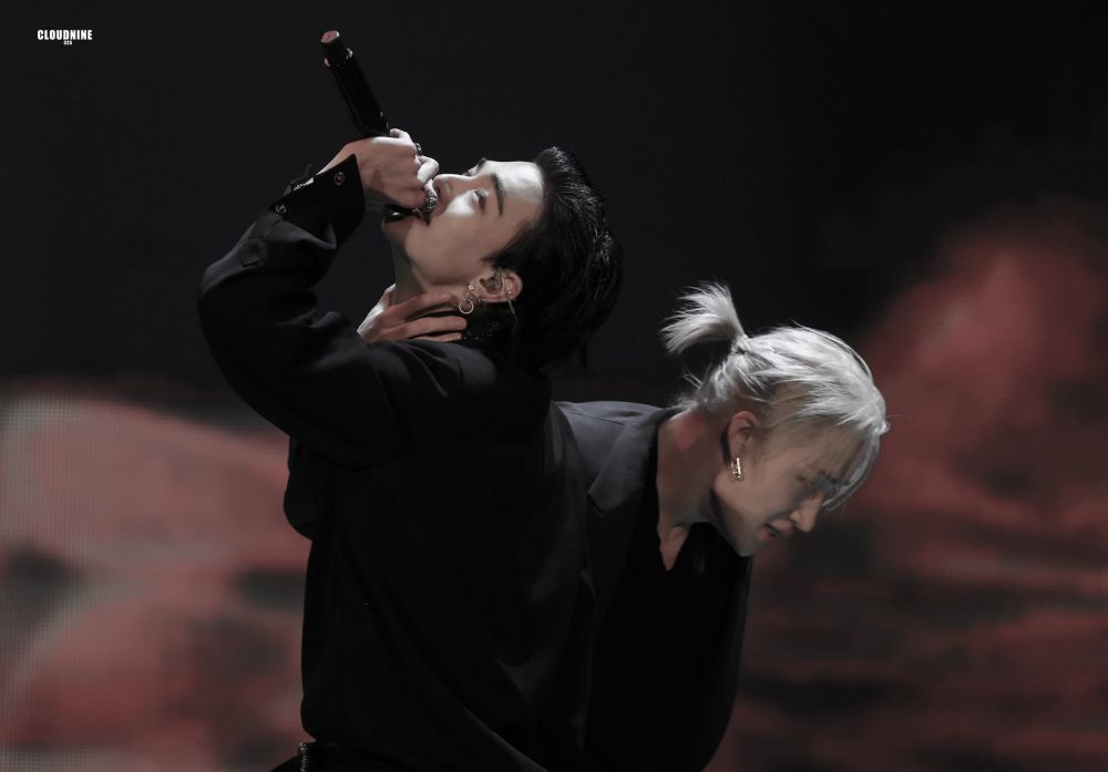 They are on stage performing escape, wearing black suits. Bang Chan has his silver hair in a ponytail and he is holding hyunjin by the neck with one hand. Hyunjin is standing straight, with his head tilted back and is singing into the mic 