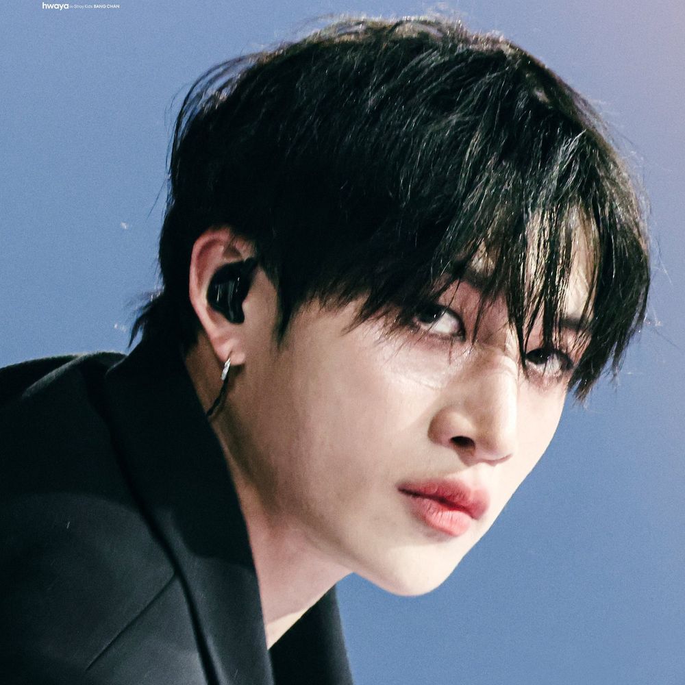 his hair is black and has strands in front of his eyes, he is wearing a black blazer, in-ears and a silver hoop earring. His face is slightly turned to the camera.