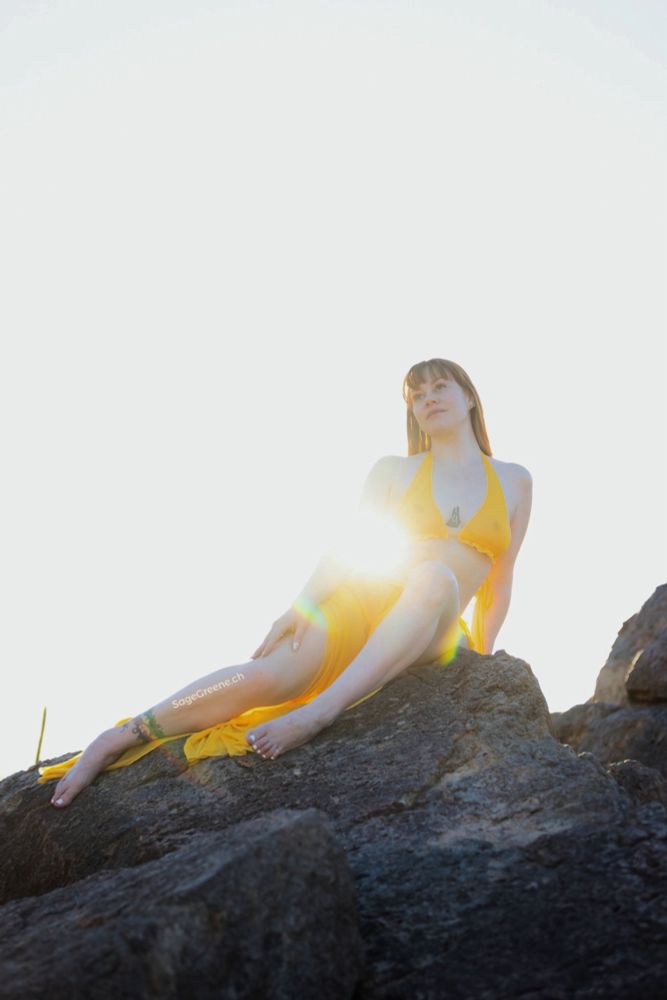 Redhead woman in sheer yellow outfit lounging on top of a large boulder little mermaid style, with the sun shining through between her arm and side.