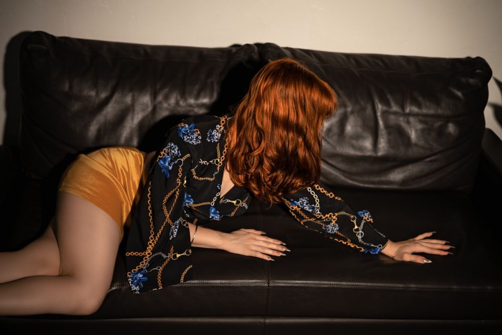 Claudia poses on a black leather couch, wearing a warm yellow mini skirt and a black shirt with blue flowers and gold chains printed on it. Her hair covers her face as she stretches out, almost crawling on the couch but in a sexy way