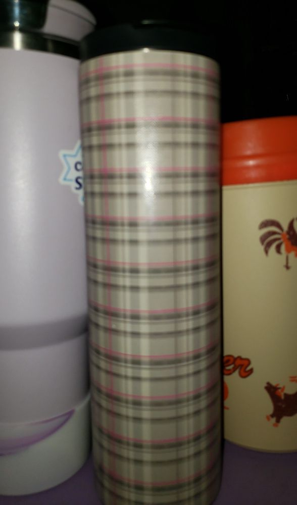 A group of three cups/thermos. The one in the center is the one currently in use. It's a homemade/fan made version of the Heaven's Dress Tartan holy water thermos Aziraphale gives to Crowley in Good Omens. There's a lens flare from the flash because I went a little heavy on the acrylic gloss sealer. It's between a lilac Stanley cup and an "iced tea jug" from Bill Miller's, a local BBQ chain. 