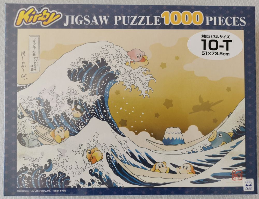 The image shows a 1000-piece jigsaw puzzle box. The puzzle design is a remake of Hokusai's "The Great Wave of Kanagawa" art with a Kirby theme. Kirby is surfing with a star on the main wave, while Waddle Dees are in boats.