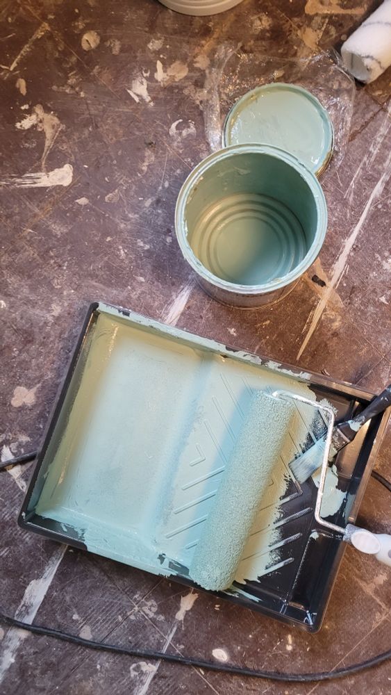 An open paint tin and a paint tray sit on the floor. The paint tray has a roller and a brush sat inside it. A very thin layer of green paint coats the inside of the tray and tin, but they are essentially dry.