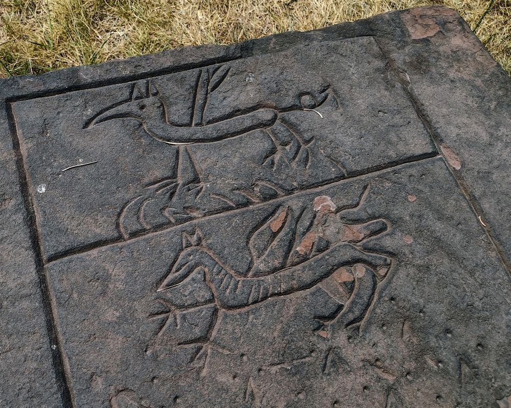 Closeup downward shot of a grayish red tabletop tomb with engravings of bizarre creatures. One looks like a cartoonish aardvark with wings and bird feet and the other similar, but with a more canine-like head and stripes.