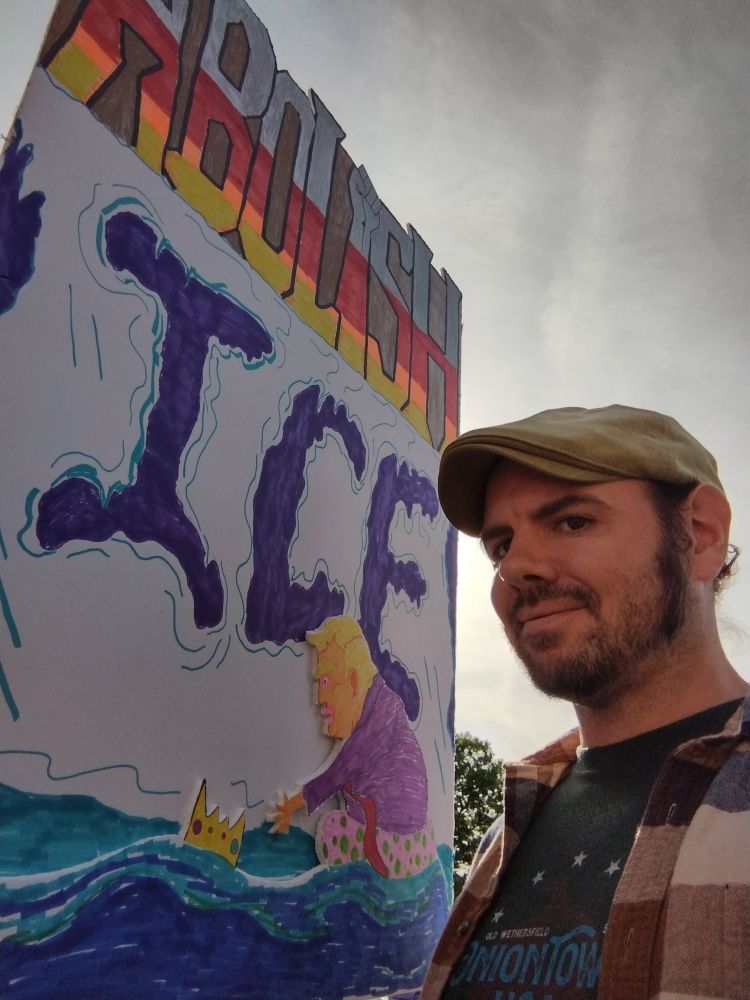 Sign that says Abolish Ice. Abolish colored in gray red orange and yellow on top of a glacier with the word ice created by melted gaps in the ice. Trump is in a pink flotation tube floating on a wave, trying to clutch a sinking crown.

Next to the sign is a man in a cap and flannel.