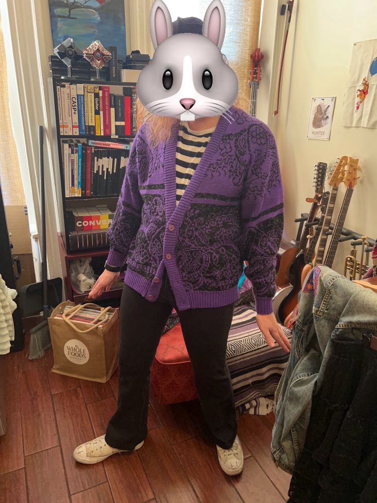 Person wearing a purple cardigan with a black floral design and a black and white striped shirt, with a cartoon rabbit emoji covering face, standing in a room with books, guitars, and a Whole Foods bag.