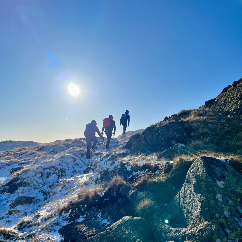 3 walkers climbing a snowy hill in the sunshine.