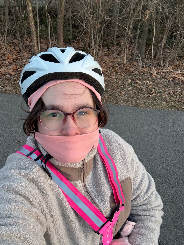 A person in a white helmet and pink light vest bundled up for winter weather. They are standing on a greenway with bare trees in the background 