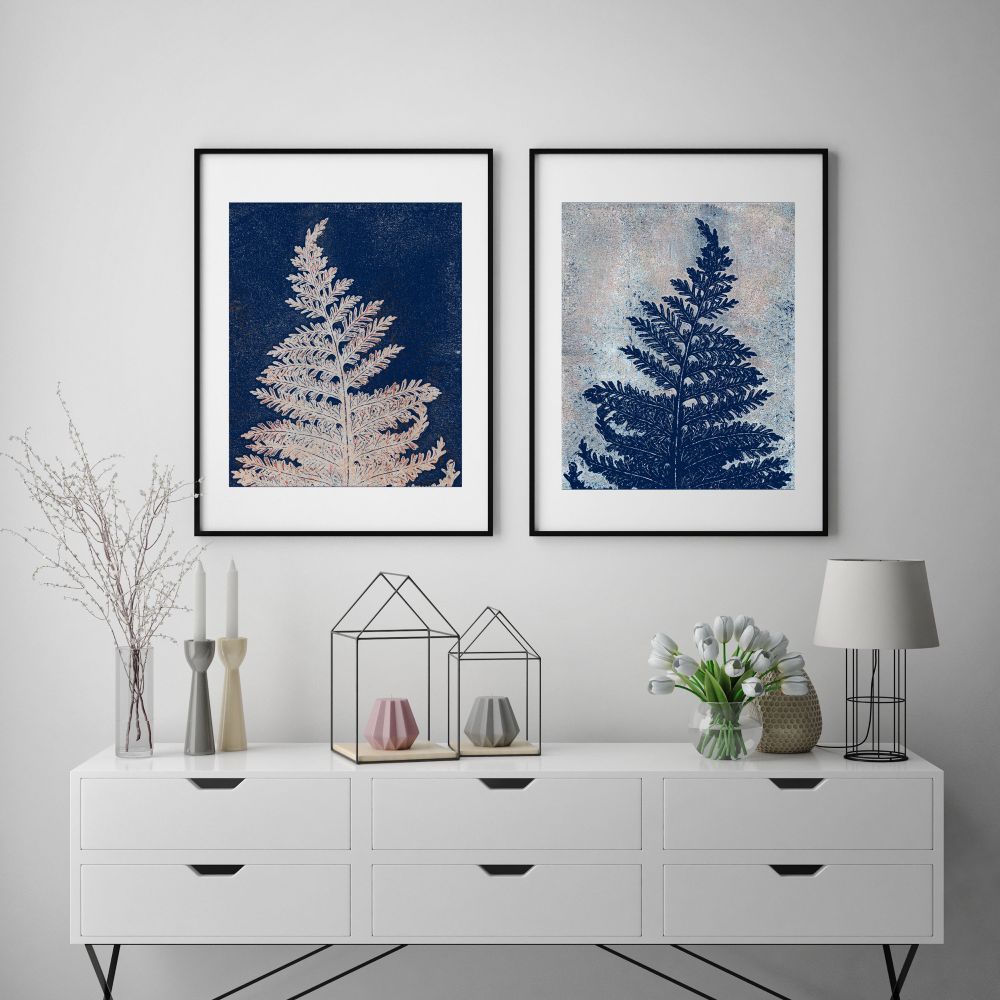 Two framed botanical art prints on a wall.  Below the prints is a set of drawers with various decorative items pleasantly displayed. Items include a lamp, a vase of white tulips and a number of candles. 