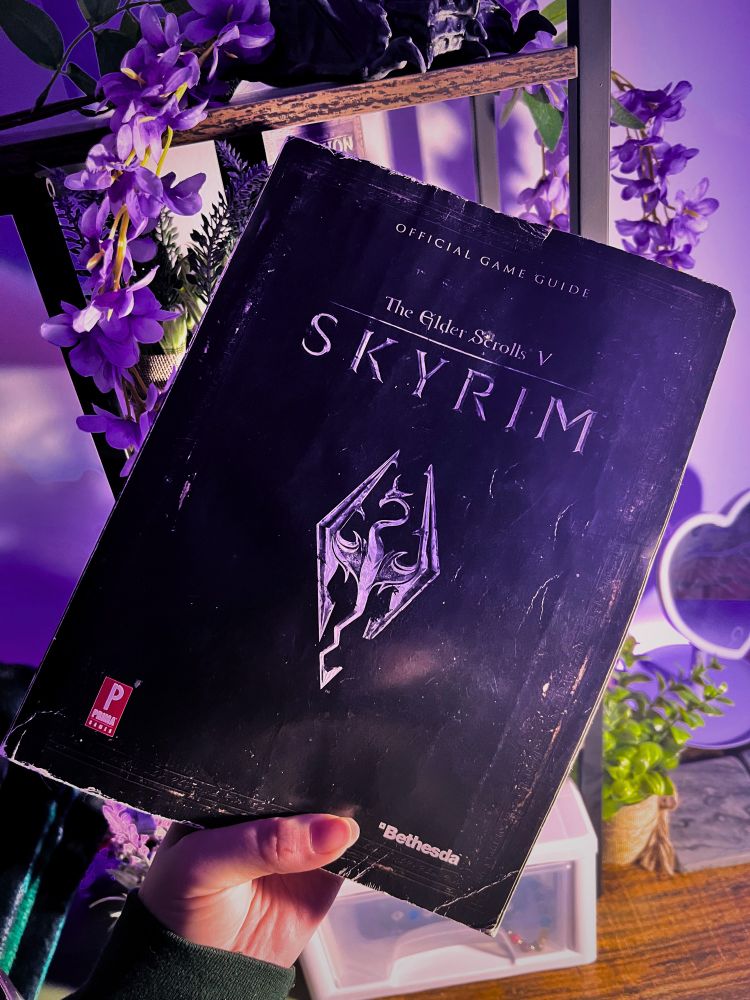 Hannah holding an original 2011 official game guide for the elder scrolls V Skyrim in front of her desk that is adorned with fake lavender and succulent plants.