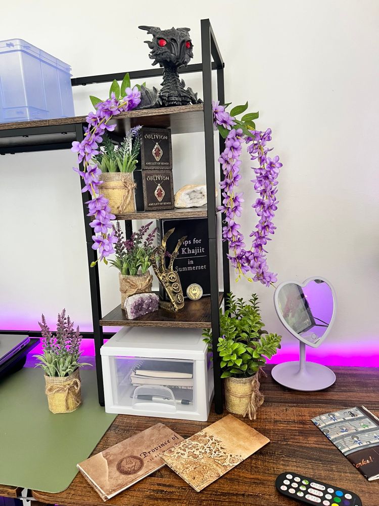 Desk with purple backlight and green desk mat. On the desk is various drawing supplies, maps of Tamriel, a fallout poster that is folded, fake wisteria, succulents, and lavender. A Tips for Khajiit faux book from ESO, a 3D printed golden claw, amethyst, quartz, two amulet of kings from oblivion, a septim coin replica, and an alduin Funko Pop (non bobble head)