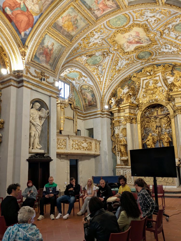 A group of adults and teens in a circle of chairs conducting a musical workshop, in a large room with frescoed vaulted ceilings and gold accents. 