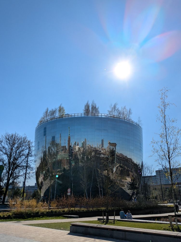 The Depot, a mirrored bowl-shaped building reflecting Rotterdam's skyline, in front of a bright blue sunny sky