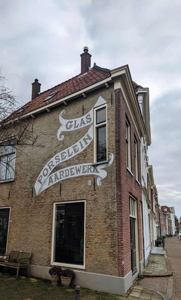 Photo depicting a brown and red brick building with a hand painted sign on its side. The sign looks like a long white scroll with the Dutch words "Glas, porselein, aardewerk" written across in uppercase letters.