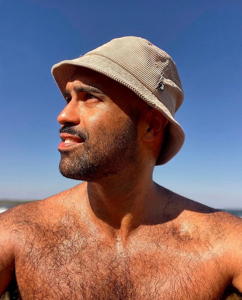 You’re truly, topless, wearing a bucket hat, at the beach, looking off to the side and not at the camera with an indigo clear sky in the background. 