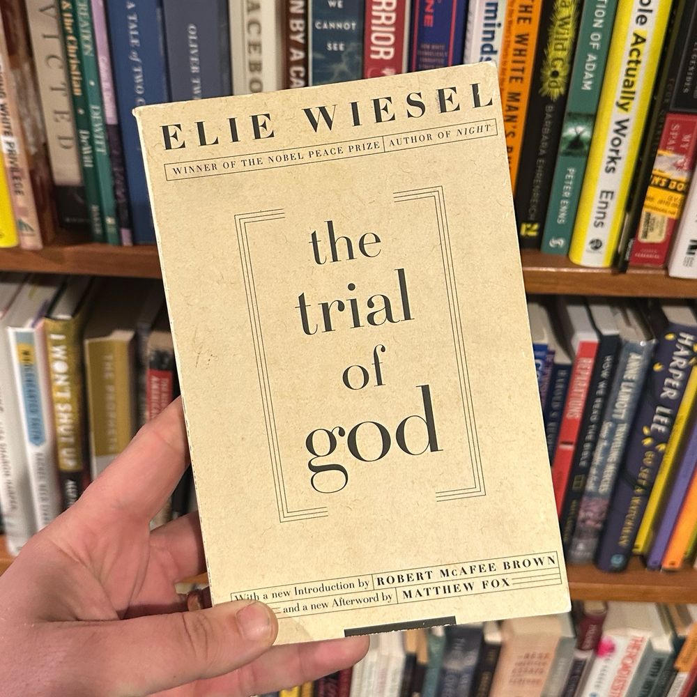 Copy of a paperback edition of The Trial of God by Ellie Wiesel held in a hand infront of a bookshelf