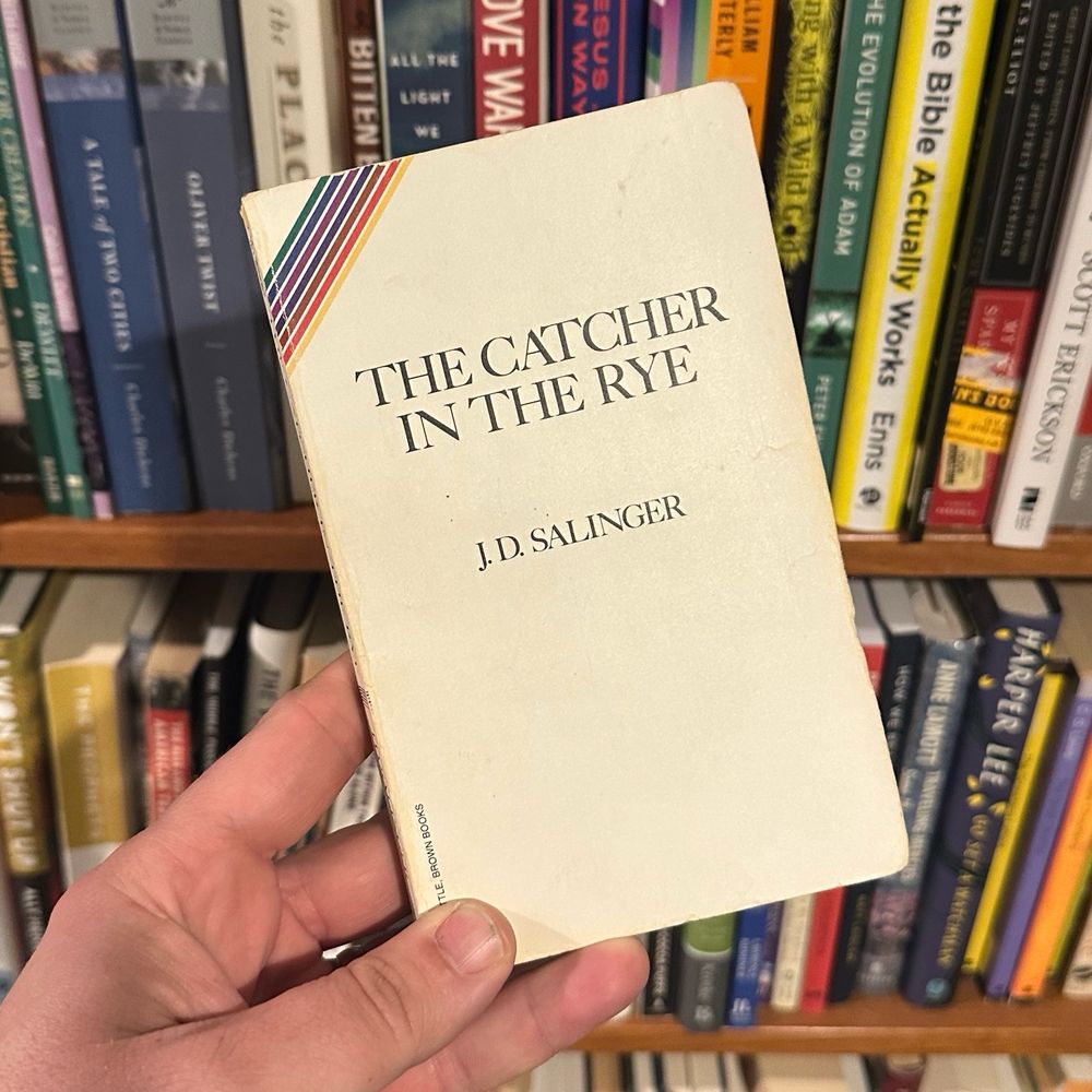 Copy of a paperback edition of The Catcher in the Rye by J.D. Salinger held in a hand infront of a bookshelf  