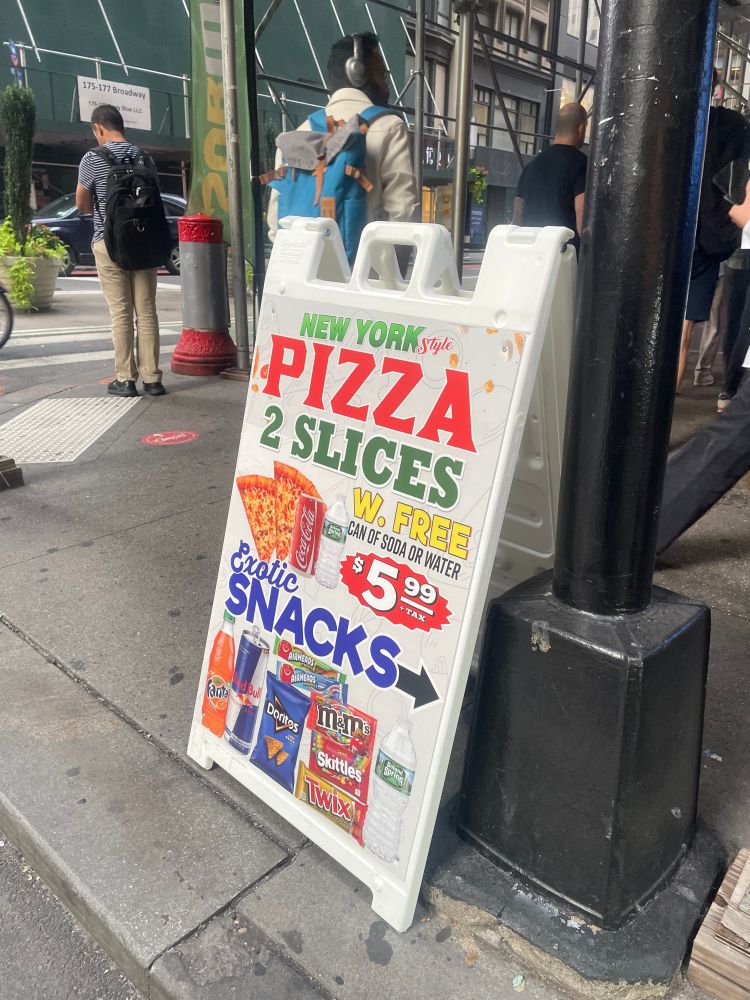 A sandwich board ad on a New York City sidewalk. It says, "New York Pizza / 2 slices / w. free can of soda or water / $5.99," with a picture of two slices of pizza with a can of Coke and a bottle of water next to it, Below, it says, "Exotic snacks" next to an arrow pointing outward. Below that, a photo of a bottle of Fanta, a can of Red Bull, Doritos, Airheads, M&Ms, Skittles, Twix, and a bottle of water.