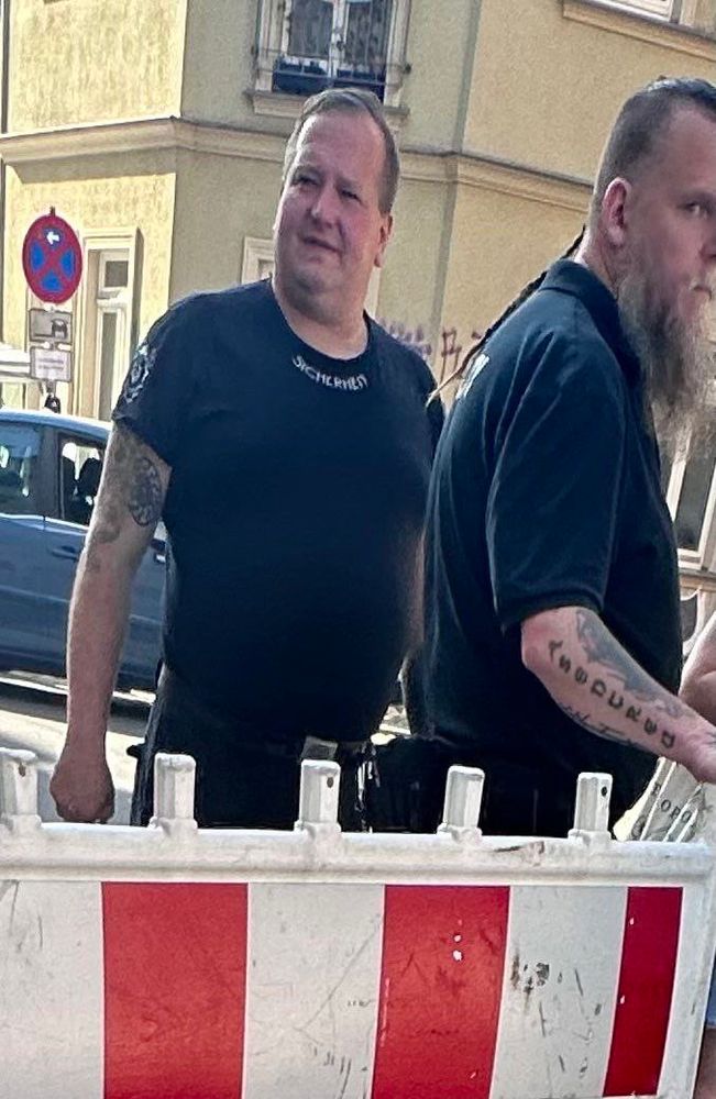 Zwei Männer stehen in der Nähe einer Baubarriere. Ein Mann hat einen tätowierten Arm und trägt ein schwarzes Hemd mit "SICHERHEIT" darauf. Der andere Mann hat einen langen Bart und ist ebenfalls schwarz gekleidet. 