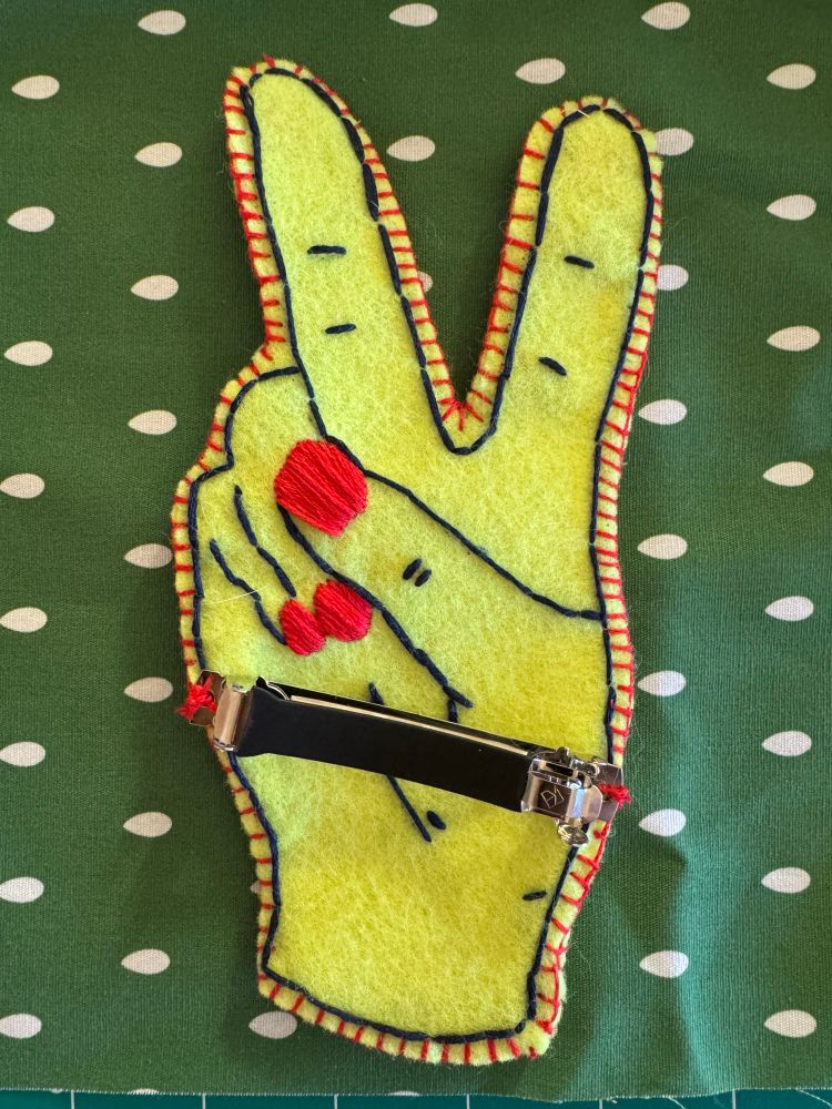 Embroidered felt — the inside of a hand making a peace sign. She’s got red nails 💅 