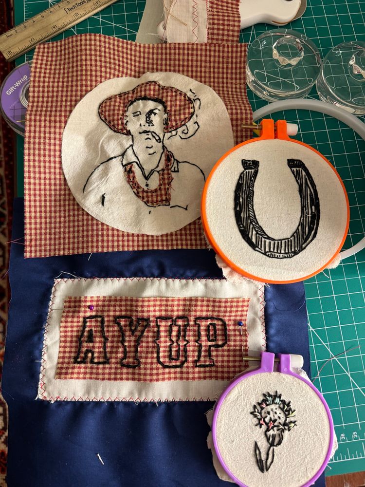 Four embroidered items: a cowboy in a red gingham bandana smoking a cigarette, a horse shoe, “ayup” embroidered on red gingham, and a thistle 