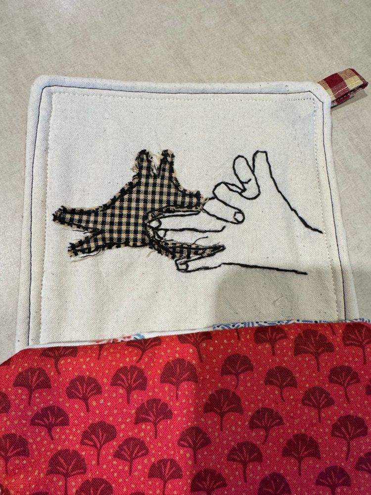 Two potholders, the top is an embroidered hand casting a dog head shadow in black gingham and the bottom is in embroidered ginkgo leaves 