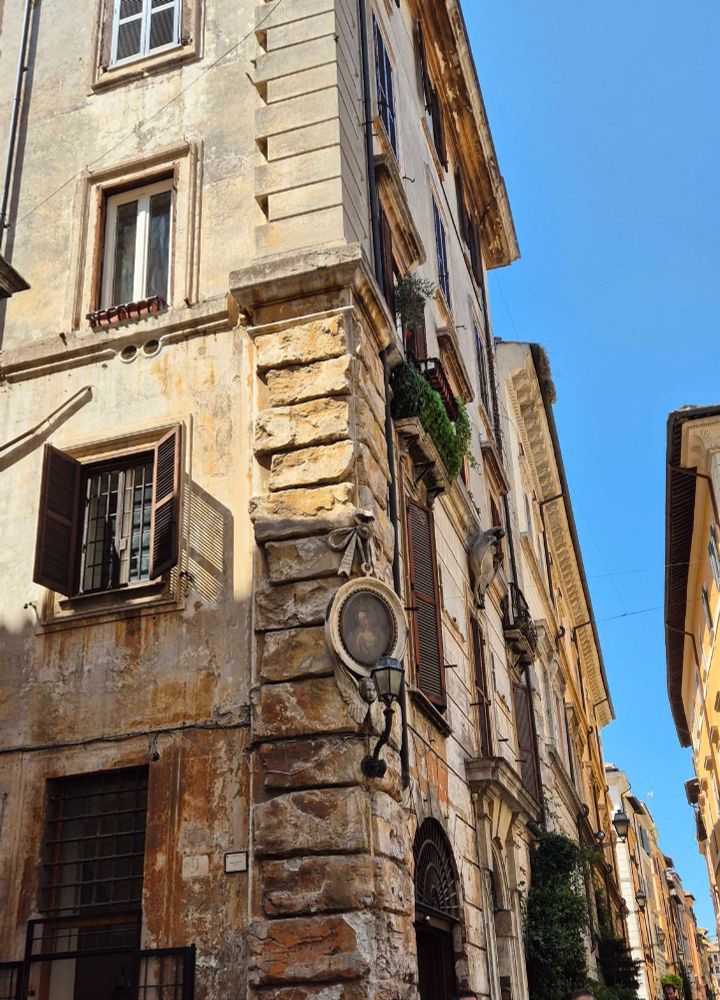 The corner of an old building in Rome, it looks very weathered. There is an oval shaped painting of a praying woman hanging on the corner of the building, a bow carved out of stone above it, which seems to depict that the painting is hanging from it. Below the painting is a carved face with angel wings. On each side of the building there's windows- some with brown shutters. It's a sunny say and the sky is bright blue.